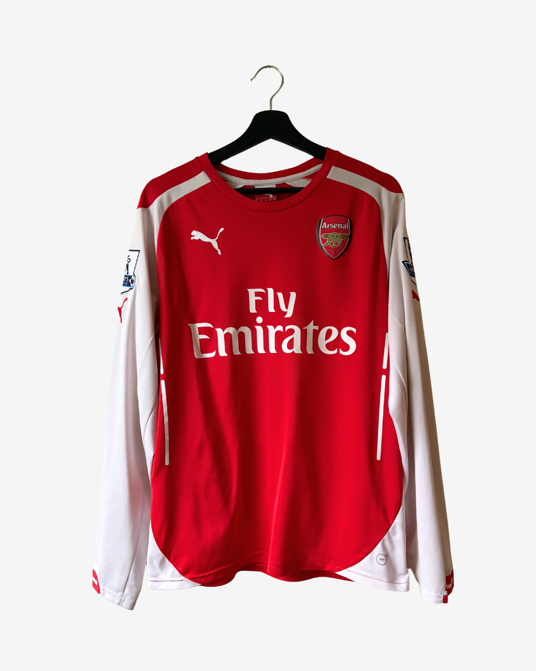 A red and white long-sleeve Arsenal football jersey by Puma, featuring the Fly Emirates sponsor logo, hanging on a black hanger against a light gray background.