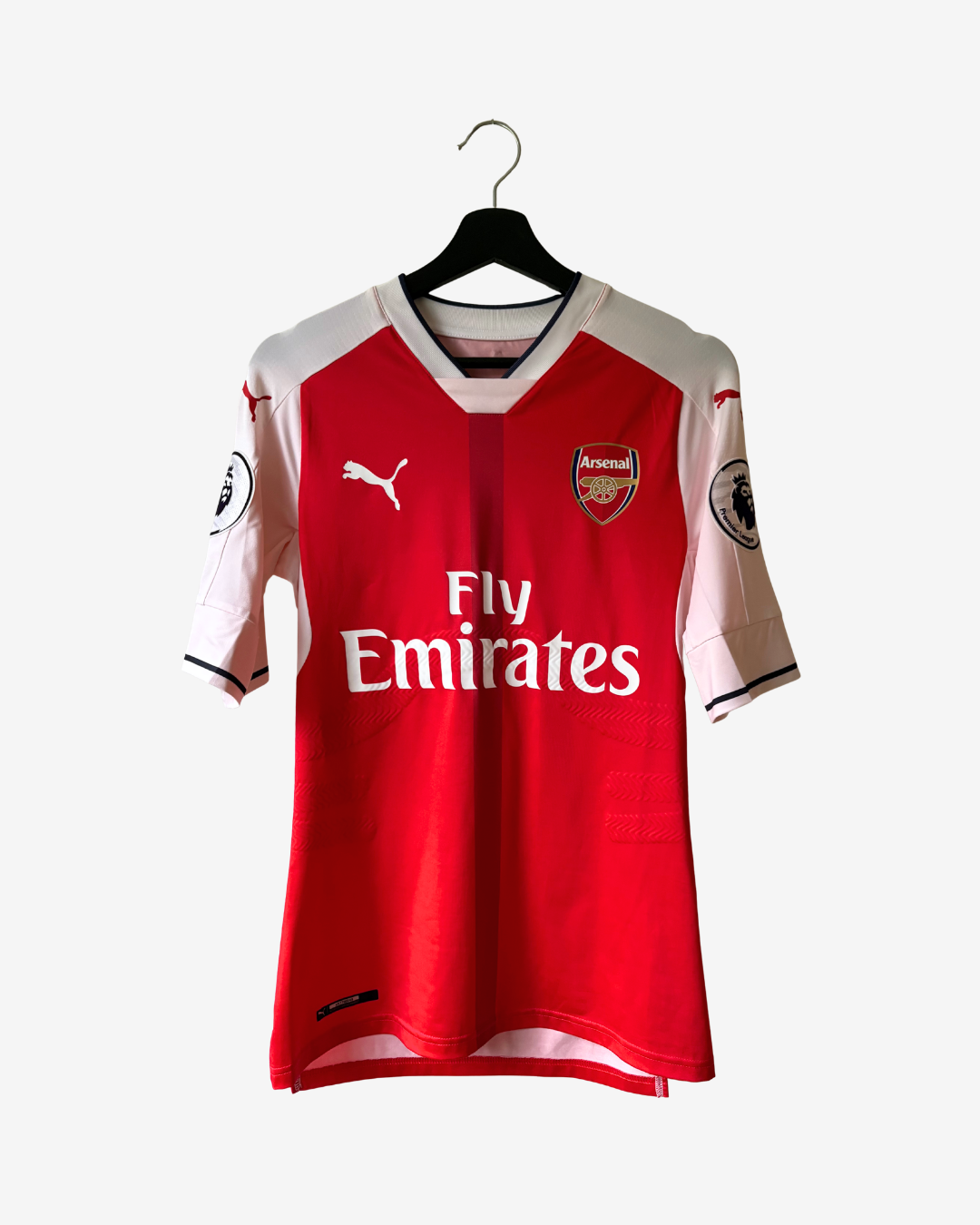 A red and white Arsenal home soccer jersey by Puma, featuring the Fly Emirates sponsor logo, hanging on a black hanger against a light gray background.