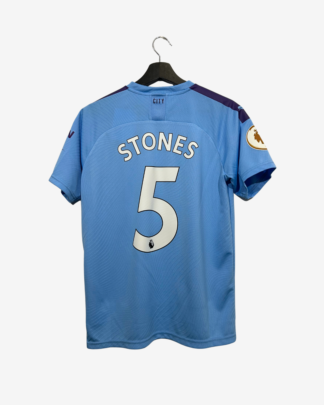 A light blue Manchester City soccer jersey shown from the back, hanging on a black hanger against a light gray background. The jersey features the name 'STONES' and number '5' in white with a black outline.