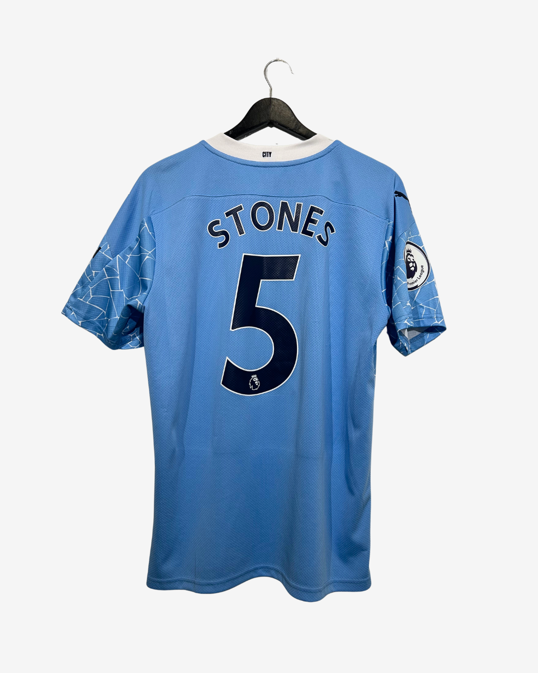 A light blue Manchester City football jersey with 'STONES 5' printed on the back, hanging on a black hanger against a light gray background.