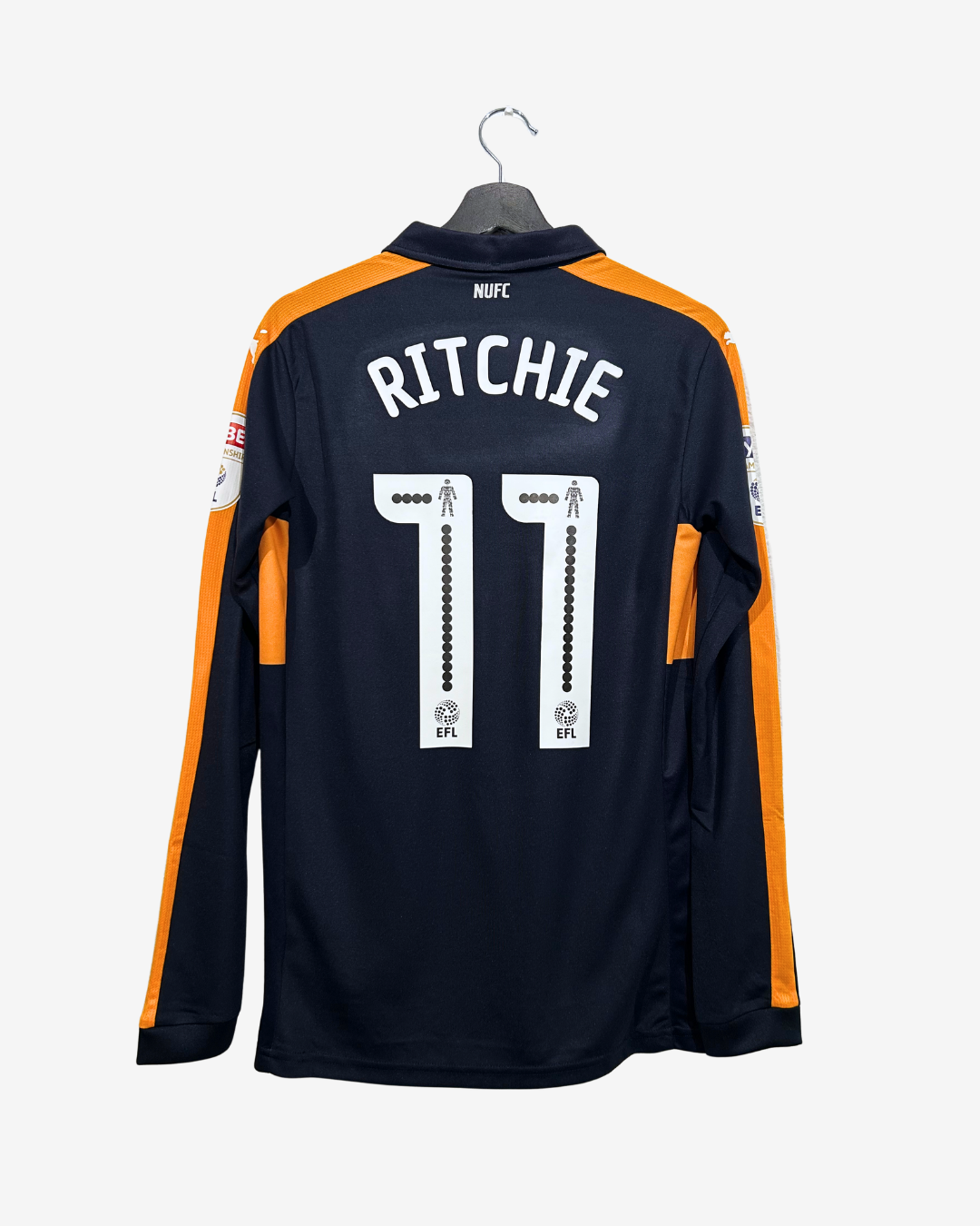Back view of a dark navy long-sleeve football jersey hanging on a black hanger against a light gray background. The jersey features orange shoulder and sleeve stripes, white 'RITCHIE' lettering and number '11' on the back, and a NUFC crest at the collar.