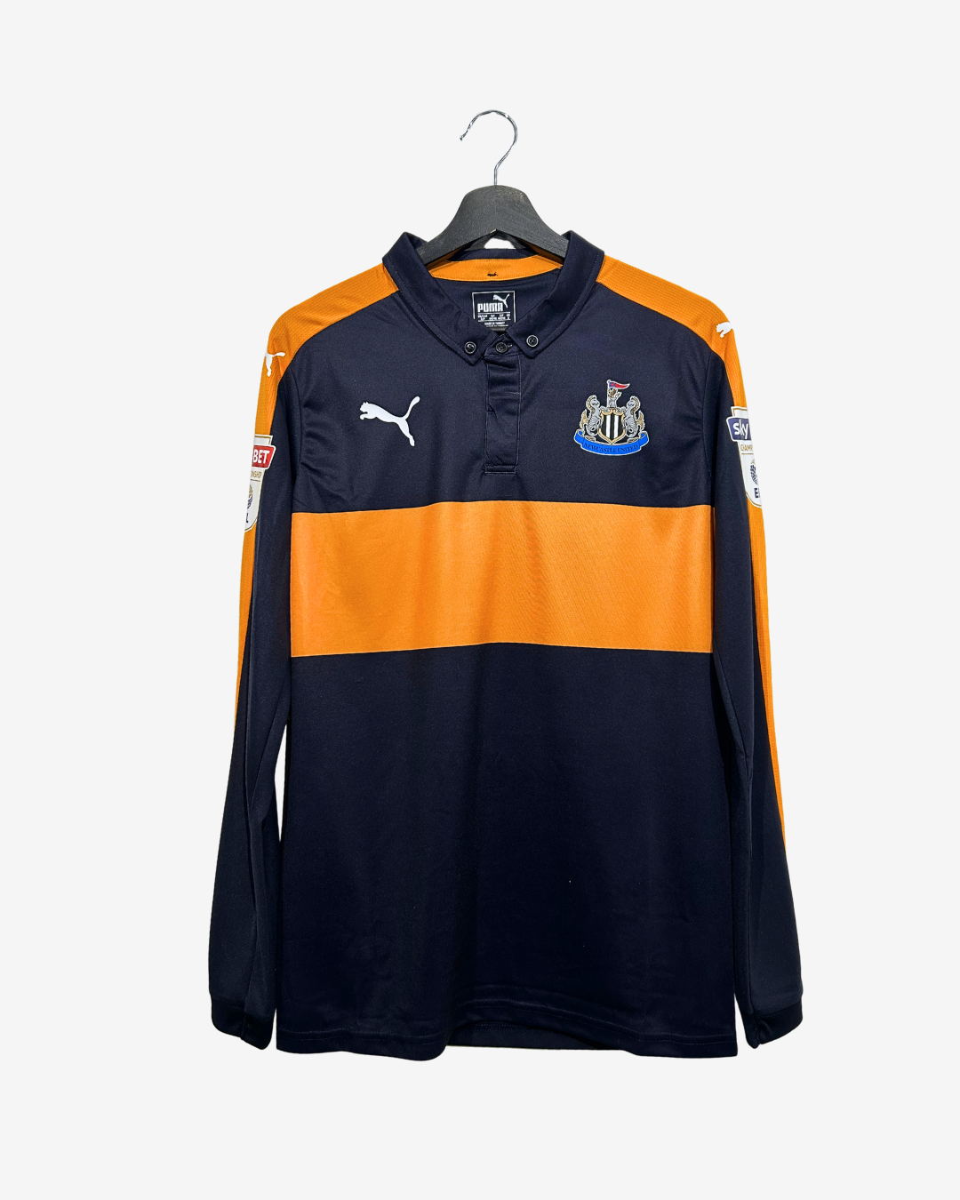A long-sleeve Puma Newcastle United football shirt in navy blue and orange, hanging on a black hanger against a light gray background.