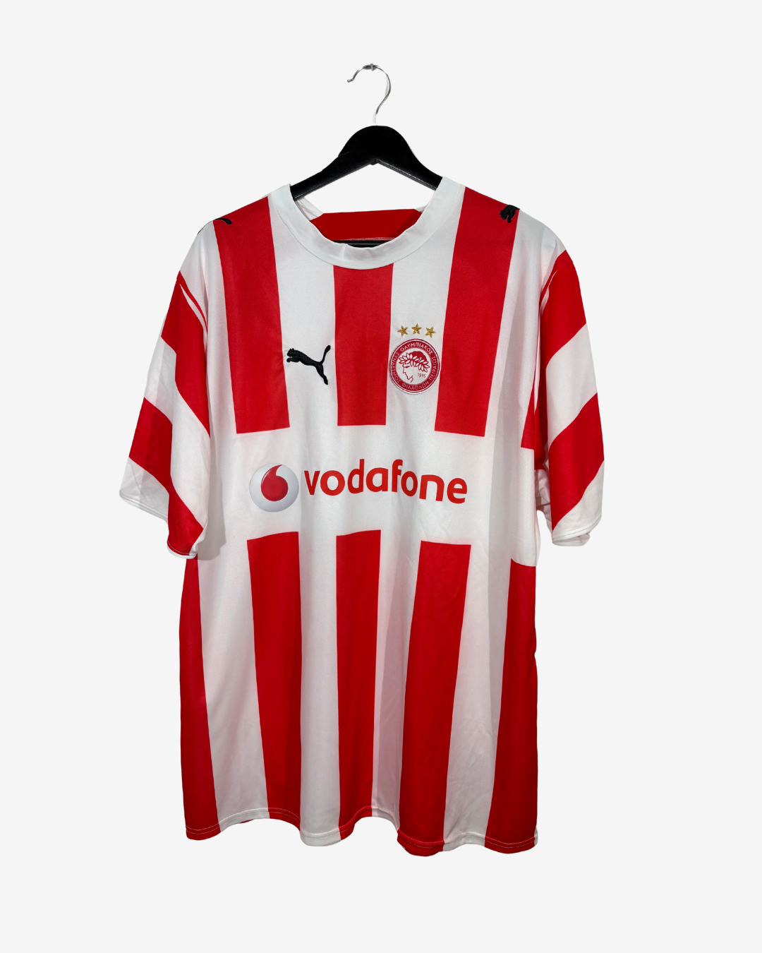 A red and white striped Puma football jersey with Vodafone sponsor logo and Olympiacos crest, hanging on a black hanger against a light gray background.