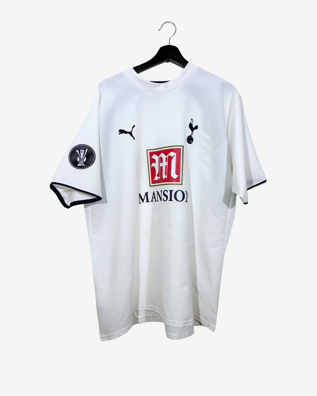 A white short-sleeve Tottenham Hotspur soccer jersey with black trim, featuring the Puma logo, the Tottenham cockerel crest, a red and white Mansions sponsor logo, and a black-and-white sleeve patch, hanging on a black hanger against a light gray background.