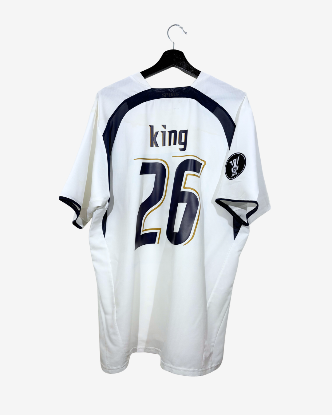 A white short-sleeve sports jersey hanging on a black hanger against a light gray background. The back of the jersey has navy blue shoulder panels, the name 'king' in navy with a gold outline, and the number '26' in large navy with a gold outline.