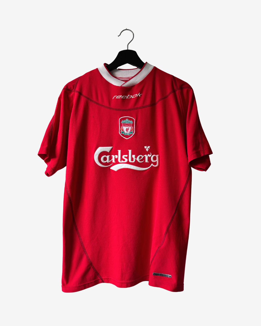 A red Reebok Liverpool football jersey with Carlsberg sponsor logo, hanging on a black hanger against a light gray background.