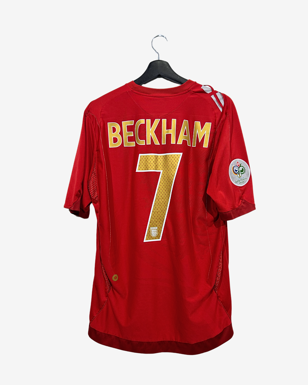 A red England national football team jersey with 'BECKHAM' and the number '7' in gold on the back, hanging on a black hanger against a light gray background.