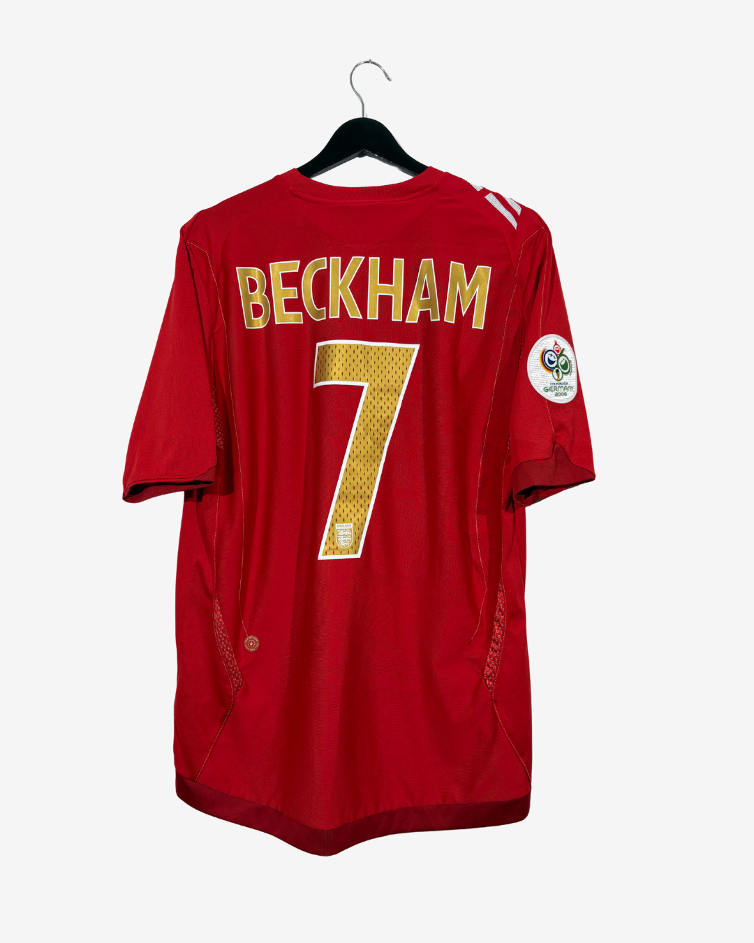 A red short-sleeve football jersey hanging on a black hanger against a light gray background. The back of the jersey features 'BECKHAM' and the number '7' in gold lettering, with a circular patch on the right sleeve.