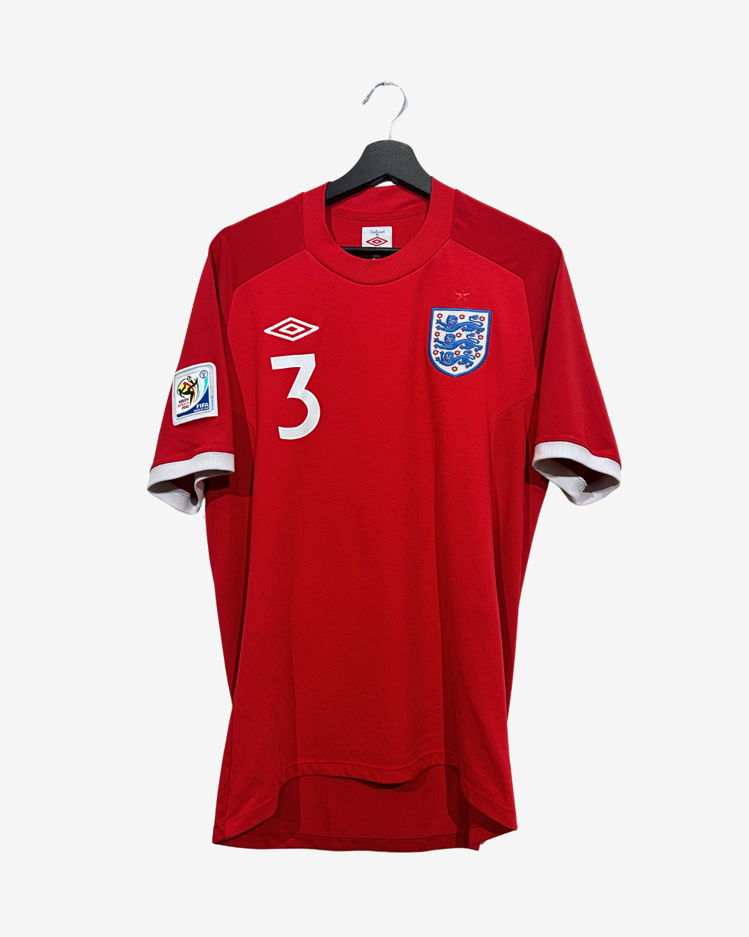 A red England national football team jersey with the number 3 on the front, featuring the Umbro logo and the England crest, hanging on a black hanger against a light gray background.