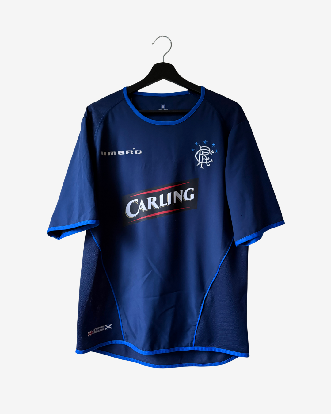 A blue short-sleeve soccer jersey hanging on a black hanger against a light gray background. The jersey features the Rangers crest on the left chest, a white 'Rangers' sponsor logo on the right chest, and a large 'CARLING' sponsor logo across the center.