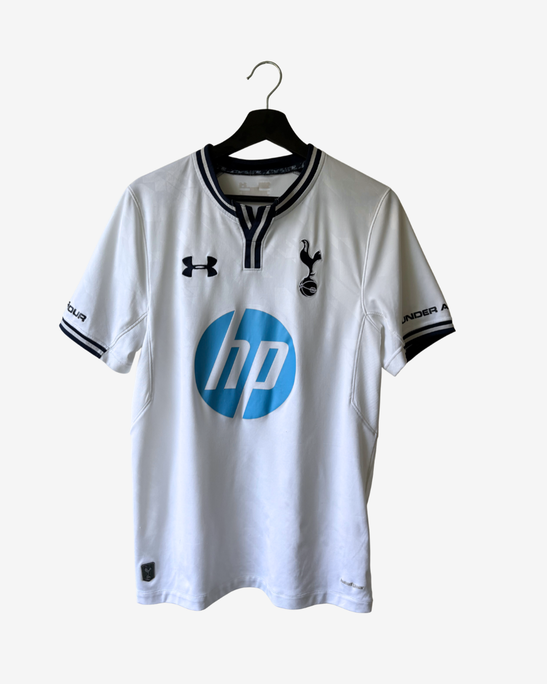 A white Tottenham Hotspur football jersey with a black and white striped V-neck collar, featuring the blue HP logo on the front, the club crest, and Under Armour branding on the sleeves, hanging on a black hanger against a light gray background.